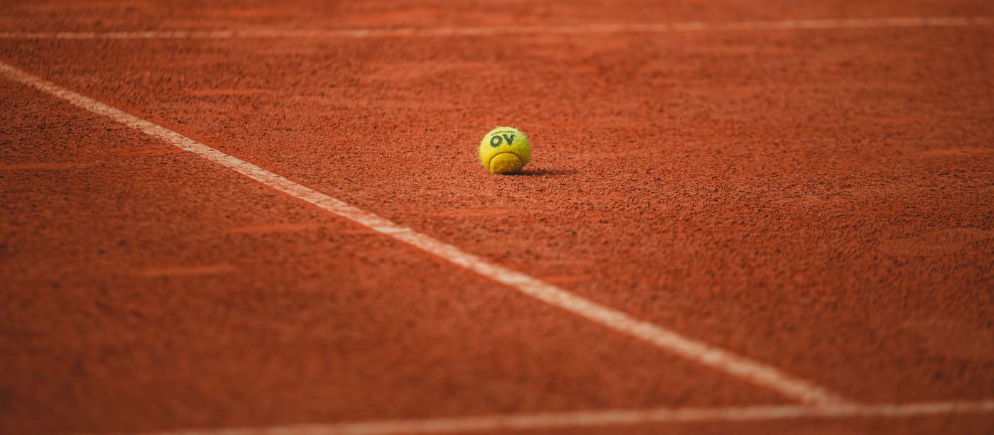 A single tennis ball resting on the red clay surface of an empty court.