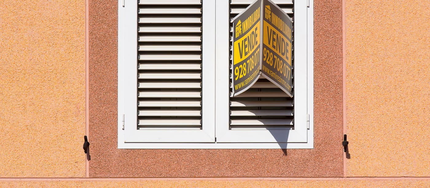 A white shuttered window on a peach-coloured building with a for-sale notice beside it.