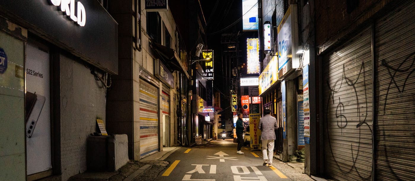 A narrow Seoul street at night, lit by shopfront signage on both sides.